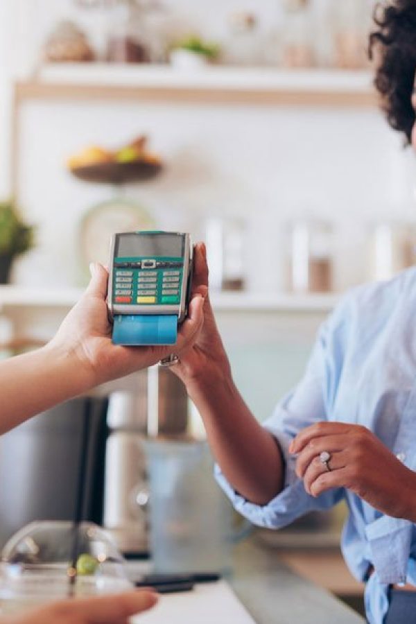 A woman holds a payment terminal while a customer makes a contactless payment in a store; promoting the Agent Program, a basket of lemons is visible in the foreground.