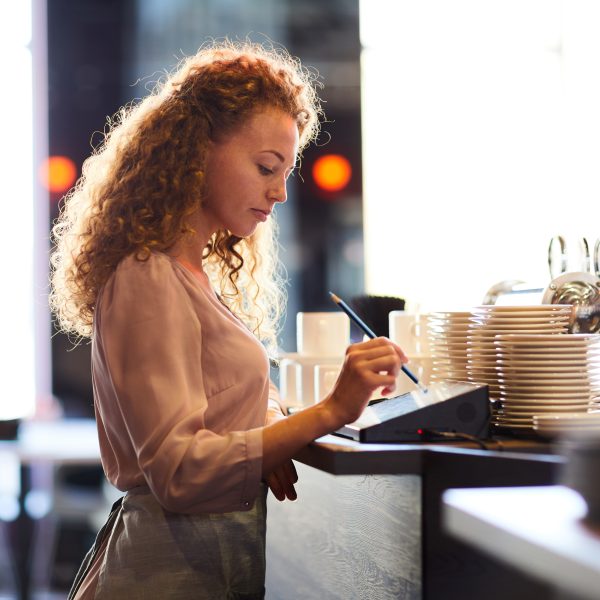 Serious pensive attractive curly-haired waitress standing at bar counter and using pencil while adding order in restaurant POS